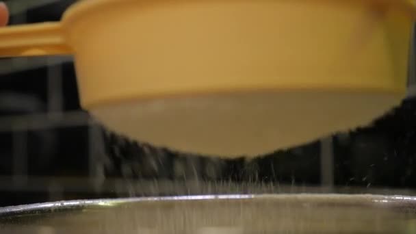 Primer plano de una fina cortina de harina de pastel blanco refinado que cae a través de un tamiz amarillo plástico en una cocina mientras se cocina una receta sobre fondo de baldosas negras, tamizando la harina en la cocina — Vídeos de Stock