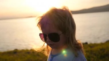Güneş gözlüklü çocuk gölde görünüyor. Sıcak yaz günü, gün batımı sırasında gölde güzel manzara. Küçük sarışın bebek altın günbatımı güneş gözlüğü ile izlerken.