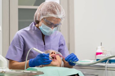 Bir dişçi, hastanın diş kalkülüsünü temizlemek için ultrason ve tükürük çıkarıcı kullanır..