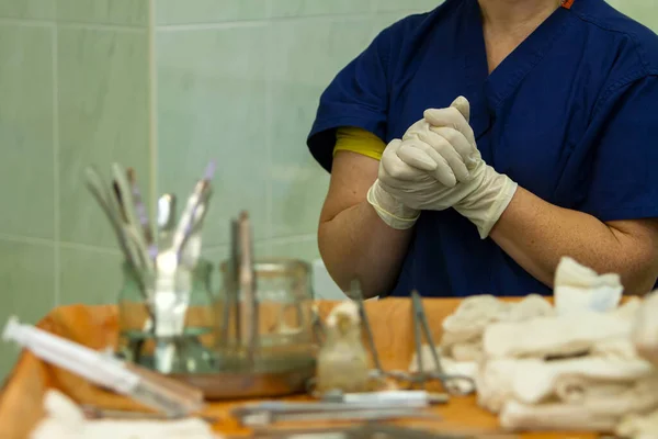 during the operation, the surgeon assistant holds his hands clasped hand in hand in front of the chest, in the foreground is a table with surgical instruments.