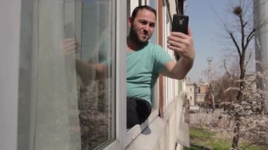 Bir adam pencere çerçevesine oturur ve akıllı telefonla iletişim kurar, konuşmayı bitirir, vedalaşır ve daireye gider. kamera hareketi pencere çerçevesinde, sağdan sola. 