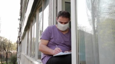 Tıbbi maskeli bir adam pencerenin kenarına oturur, İbranice bir defterde kalemle yazar, sağdan sola, sokaktan statik bir çekim. Tam HD, ses yok..