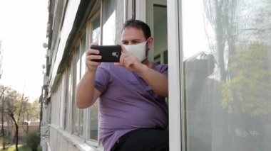 Yüzünde tıbbi maske olan bir adam pencere çerçevesinde oturur ve akıllı telefondan video çeker. Sokak manzaralı, orta ölçekli, full HD, ses yok..