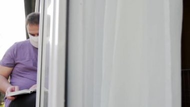 Tıbbi maskeli bir adam pencere kenarına oturur, bir kitap okur, sağ eliyle sayfayı çevirir. Kamera pencerenin çerçevesinden sağa sola hareket ediyor. Orta çekim, Full HD, ses yok.