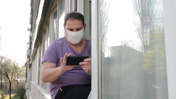 一个满脸面具的男人坐在窗框上 用智能手机在感情上玩游戏 举起拳头庆祝胜利 然后站起来离开 摄影机从一条街移动到另一条街 — 图库视频影像