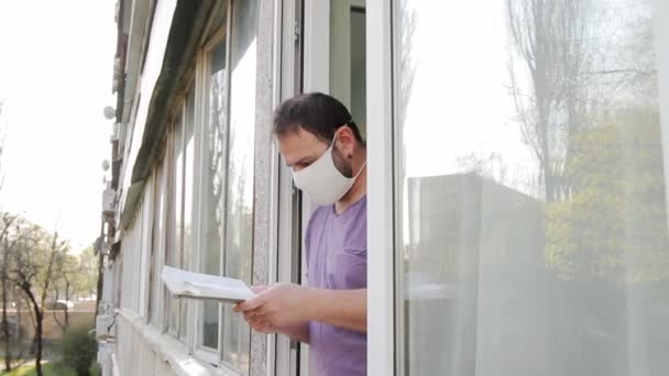 医療用マスクの男が本を手に開いた窓に近づき 本を読み 右の手でページをめくる カメラの動きが通りからアパートに向かって左から右へ — ストック動画