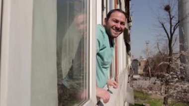 Bir adam açık bir pencerenin yanında duruyor, derin bir nefes alıyor, gülümsüyor ve neşeyle kenarlara bakıyor. Pencere çerçevesindeki kamera hareketleri daireden sokağa soldan sağa doğru. orta çekim