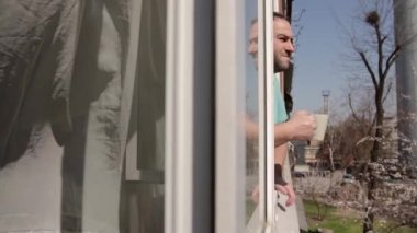 Sağ elinde bej bir bardakla açık bir pencerede bir adam beliriyor, bardaktan su içiyor ve etrafa bakıyor. kamera hareketi pencere çerçevesinde soldan sağa hareket ediyor.