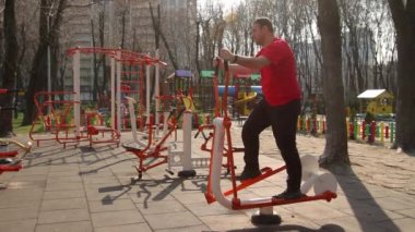 Orta yaşlı bir adam sokak ortasında spor salonunda spor yapıyor. Kardiyo simülatöründe, öğleden sonra, uzak çekim, full HD, ses yok.
