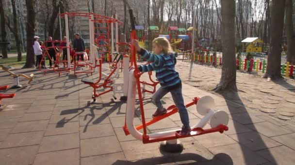 乌克兰 2020年4月5日 一个6岁的女孩在一个公共运动场上 在一个有氧模拟器上做运动 拍了一个长镜头 一群人在后面聊天 没有声音 — 图库视频影像
