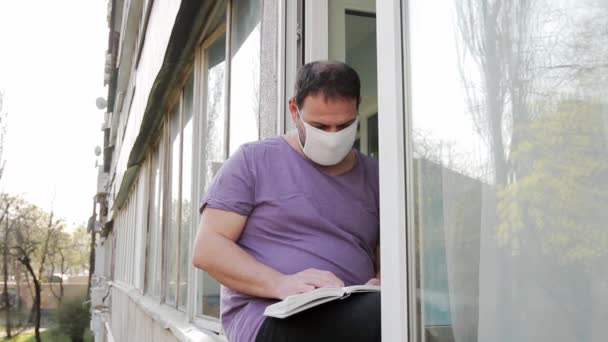 医療マスクの男が窓枠の上に座り 本を読み 右手でページをめくる ミディアムショット フルHd サウンドなし — ストック動画