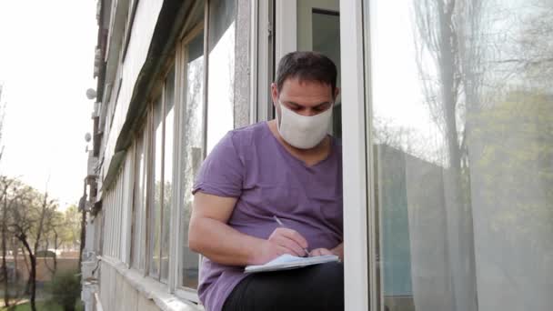 Man Medicinsk Mask Sitter Fönsterkarm Skriver Avsiktligt Med Penna Anteckningsbok — Stockvideo