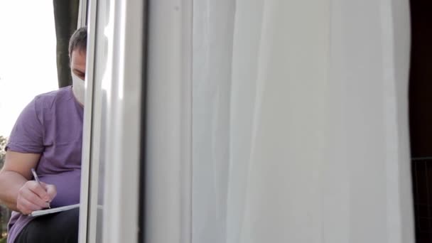 Homme Portant Masque Médical Assoit Sur Cadre Fenêtre Écrit Intensément — Video