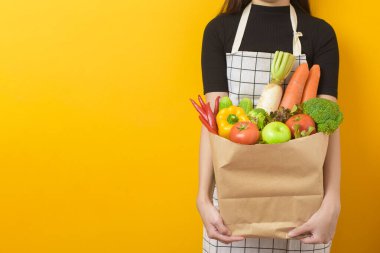  Stüdyo sarı arka planda sebzeleri market poşetinde tutan güzel bir genç kadın var. 