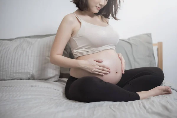 美しいですアジアの妊娠中の女性座っていますベッドの上に触れます彼女の腹 — ストック写真