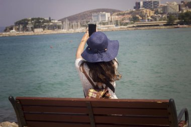 Deniz kenarındaki bir bankta oturan kadınlar, turistlerin özçekimlerini çekiyor.