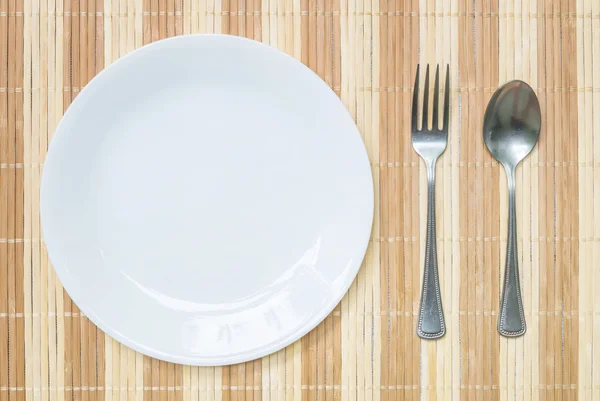 Plato cerámico blanco de primer plano con tenedor inoxidable y cuchara sobre alfombra de madera fondo texturizado sobre mesa de comedor en vista superior — Foto de Stock