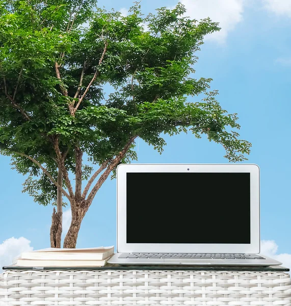 Computadora de primer plano en madera borrosa tejer mesa y libro en árbol y cielo azul con fondo de nube, hermoso lugar de trabajo en el concepto al aire libre — Foto de Stock
