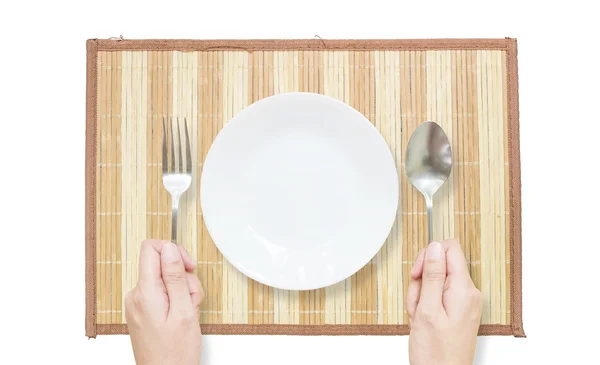 Plato cerámico blanco de primer plano con tenedor inoxidable y cuchara en mano de mujer sobre estera de madera marrón aislada sobre fondo blanco en vista superior — Foto de Stock