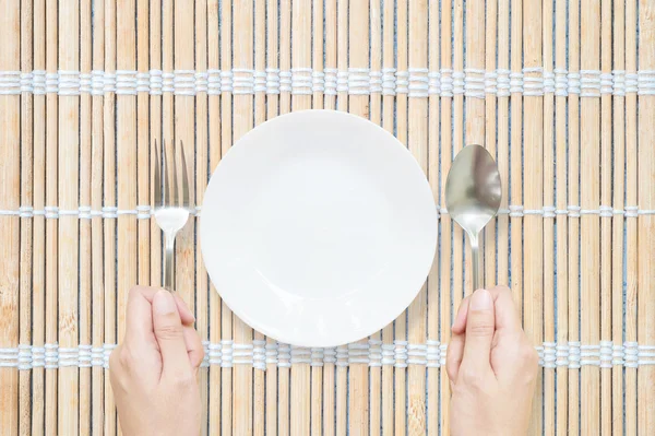 Närbild vit keramisk maträtt med rostfritt gaffel och sked i kvinnan hand på trä matta texturerad bakgrund på matbordet i topp vy — Stockfoto