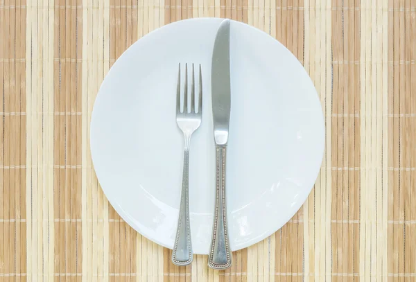 Primer plano plato de cerámica blanca con tenedor inoxidable y cuchillo en la estera de madera fondo texturizado en la mesa de comedor en la vista superior en la comida es un concepto muy delicioso — Foto de Stock