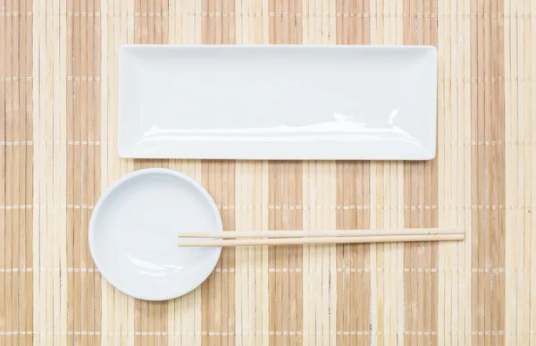 Primer plano plato cuadrado de cerámica blanca y cáliz con palillos de madera en la estera de madera fondo texturizado en la mesa de comedor en vista superior — Foto de Stock