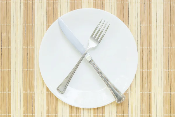 Closeup white ceramic dish with stainless fork and knife on wood mat textured background on dining table in top view in unfinished eating concept — Stock Photo, Image