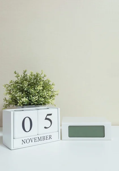 Άσπρο ξύλινες ημερολόγιο με μαύρο 5 Νοεμβρίου λέξη με ρολόι και φυτών σε λευκό ξύλινο γραφείο και κρέμα ταπετσαρία υφή φόντου, επιλεκτική εστίαση στο ημερολόγιο — Φωτογραφία Αρχείου