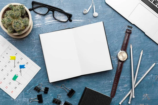 Opened notebook and other office equipment such as computer, pencil, and glasses on wooden office desk.