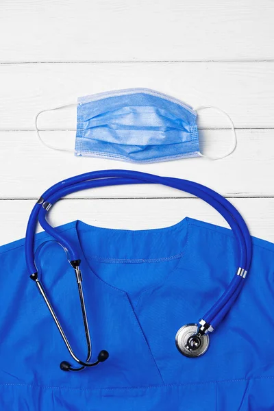 白い木製の背景に聴診器とフェイスマスクと医師の医療制服 — ストック写真