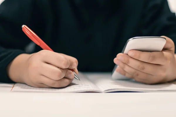 Primer plano de un joven estudiante con un smartphone escribiendo en un cuaderno . —  Fotos de Stock