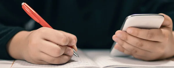 Primer plano de un joven estudiante con un smartphone escribiendo en un cuaderno . —  Fotos de Stock