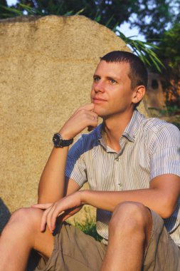 Rahat çekici gülümseyen adam oturup bir gömlek, şort ve büyük bir taş yakınındaki saat
