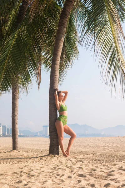 Сексуальная загорелая девушка в солнечных очках возле пальмы на пляже в зеленом трикотажном купальнике — стоковое фото