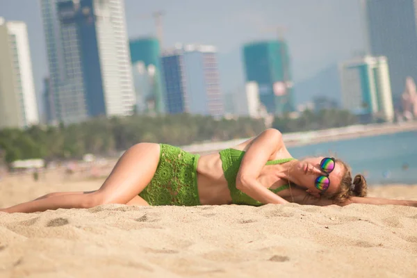 Сексуальная загорелая девушка в солнечных очках возле пальмы на пляже в зеленом трикотажном купальнике, лежит на песке — стоковое фото