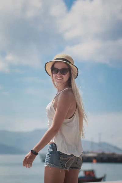 Красивая женщина стоит на городской набережной и наслаждается видом, в соломенной шляпе, спиной к камере — стоковое фото