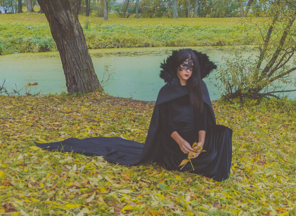 Beautiful girl in a black mask and gown is knee collects leaves.