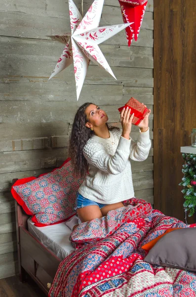 Chica en suéter blanco desempaca regalo de Navidad en la cama . — Foto de Stock