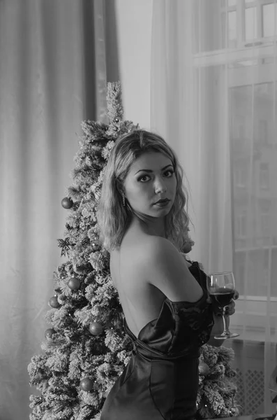 Schwarz ist weiß, Nacht am Weihnachtsbaum posiert ein pikantes Mädchen im Abendkleid mit offenem Rücken und einem Glas Wein — Stockfoto