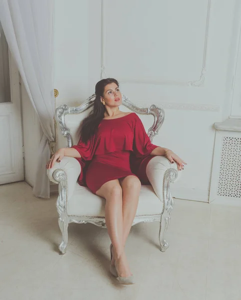 Menina bonita em vestido vermelho posando sentado em uma cadeira vintage . — Fotografia de Stock