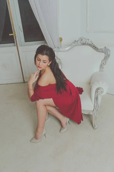 Esponendo la spalla siede in una sedia vintage, posa ragazza attraente in un abito rosso — Foto Stock