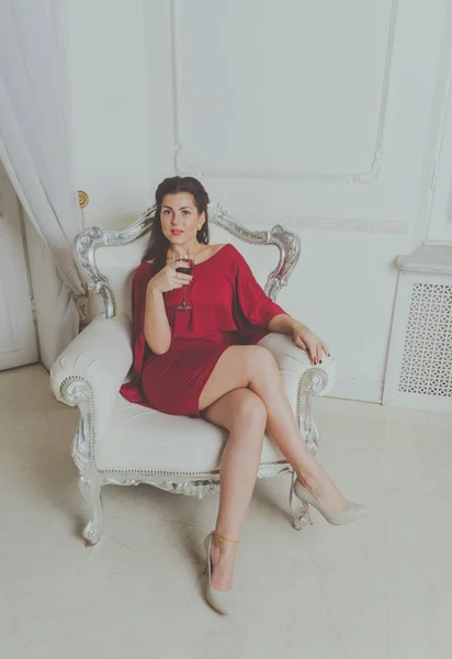 Z kieliszkiem wina siedzi na krześle vintage, pozowanie atrakcyjny dziewczynka w czerwonej sukience — Zdjęcie stockowe