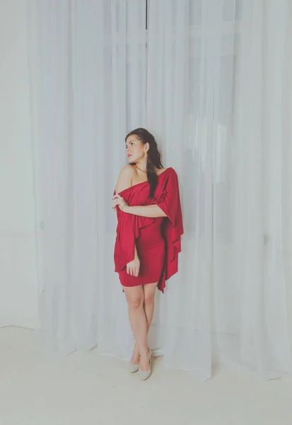 Παθιασμένος κορίτσι με κόκκινο φόρεμα στέκεται στο παράθυρο — Φωτογραφία Αρχείου