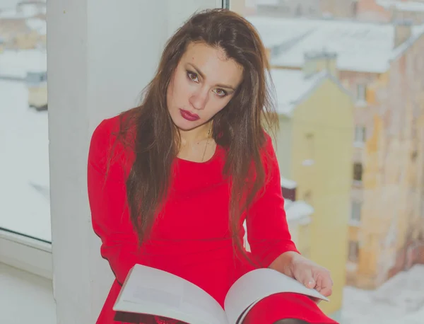 Menina em vermelho com um livro na mão sentado à janela, olhando para a câmera — Fotografia de Stock