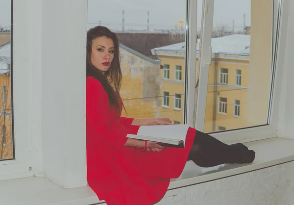 Olha para a câmera, sentado na janela com um livro uma menina bonita em um vestido — Fotografia de Stock