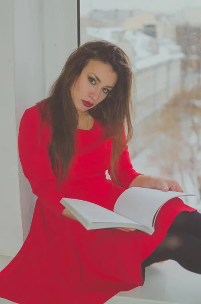 Sentado na janela com um livro uma menina bonita em um vestido — Fotografia de Stock