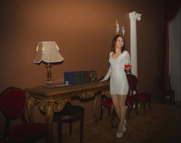 Chica en un vestido blanco con una copa de vino en el vestíbulo cerca de la mesa con el teléfono — Foto de Stock