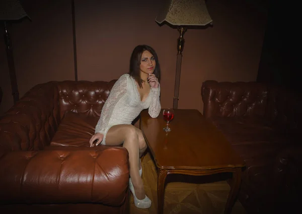 Flicka i en vit klänning med ett glas vin sitter på soffan — Stockfoto