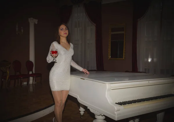 Niña en un vestido blanco con una copa de vino de pie cerca del piano —  Fotos de Stock