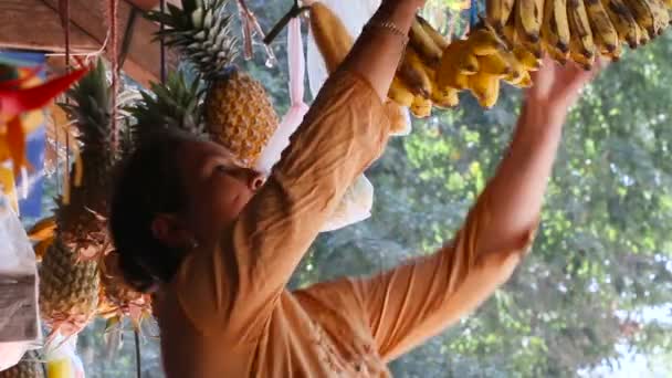 SIEMREAP, CAMBODIA - 8 de março de 2013. Loja de rua com frutas penduradas para venda abacaxi, banana, melancia. A mulher vende bananas frescas. Camboja . — Vídeo de Stock